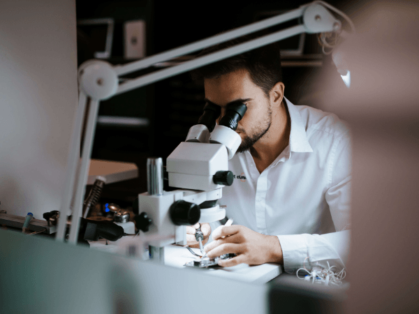 Férfi fehér ingben egy iPhone-szerviz mikroszkópján vizsgálja a telefon alkatrészét.