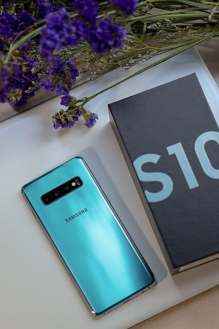 Egy asztalon lila virág mellett egy kék Samsung Galaxy S10 készülék, mellette a gyári dobozával.